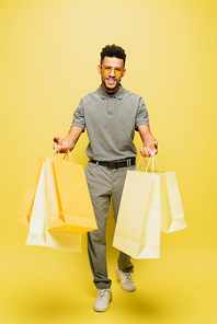 full length of cheerful african american man in sunglasses and grey tennis shirt holding shopping bags on yellow