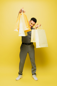 full length of positive african american man in sunglasses and grey tennis shirt holding shopping bags on yellow