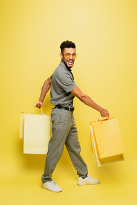 full length of happy african american man in sunglasses and grey tennis shirt standing with shopping bags on yellow