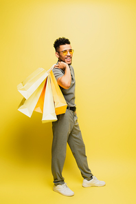 full length of joyful african american man in sunglasses and grey tennis shirt holding shopping bags on yellow