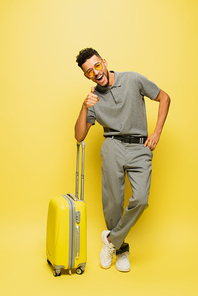 full length of happy african american man in sunglasses and grey tennis shirt showing thumb up near luggage on yellow