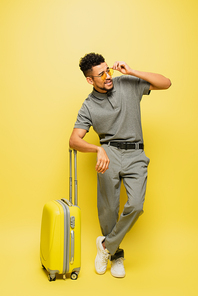 full length of stylish african american man in sunglasses and grey tennis shirt standing near luggage on yellow