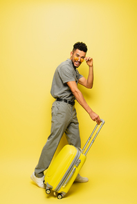 full length of cheerful young african american man adjusting sunglasses and standing with luggage on yellow