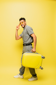 full length of happy african american man in sunglasses and grey tennis shirt carrying luggage on yellow