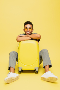 full length of cheerful african american man in sunglasses sitting with luggage on yellow