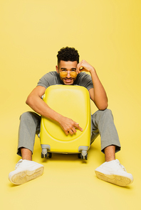 full length of positive african american man in sunglasses sitting with luggage on yellow