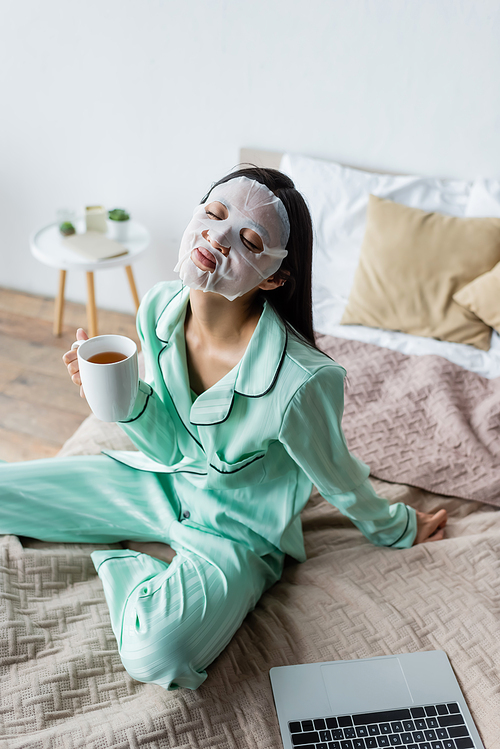 asian woman in sheet mask sitting on bed with cup of tea and closed eyes near laptop