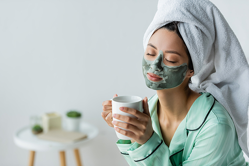 pleased asian woman in clay mask, pajamas and towel on head holding cup of tea with closed eyes