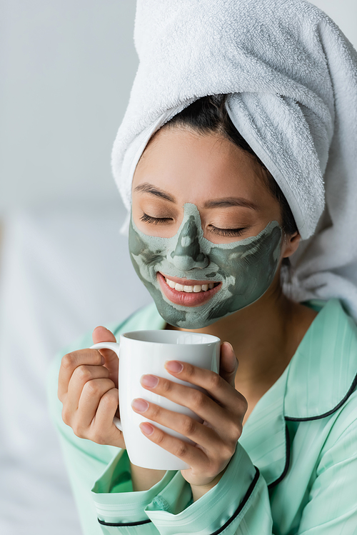 joyful asian woman with clay mask on face and towel on head drinking tea with closed eyes