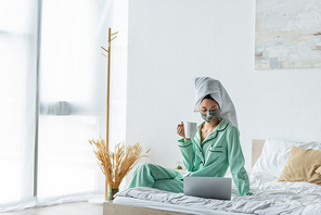 asian woman in clay mask and pajamas sitting on bed with cup of tea and looking at laptop