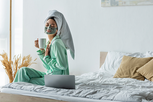 pleased asian woman with closed eyes holding cup of tea while sitting near laptop in bedroom