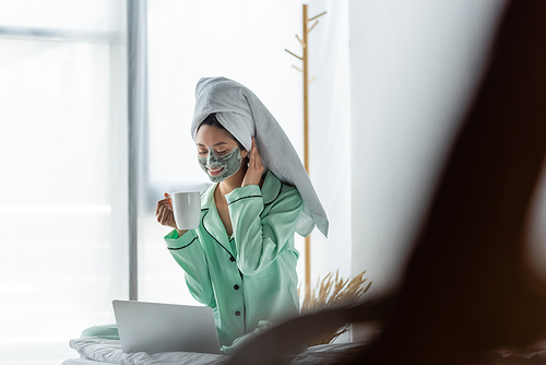 smiling asian woman in pajamas and clay mask sitting with cup of tea near laptop in bedroom