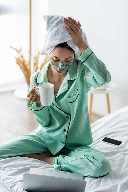 asian woman in clay mask and towel on head sitting on bed with cup of tea near laptop