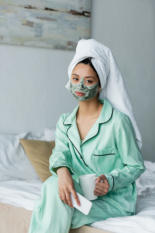 asian woman in pajamas and clay mask sitting on bed with smartphone and cup of tea