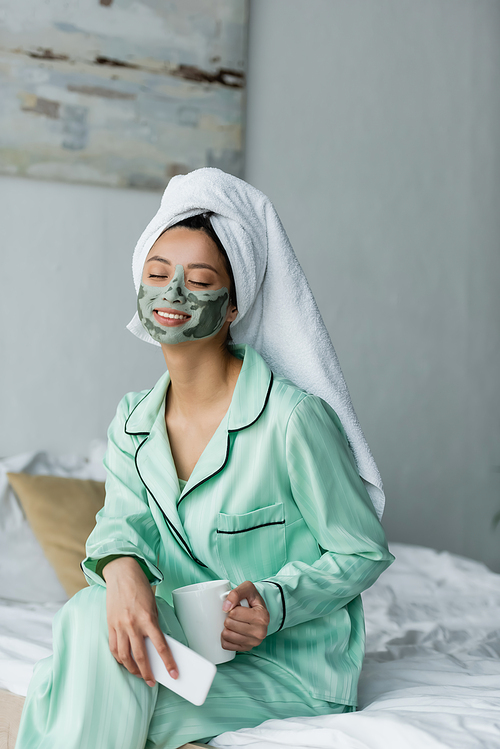 pleased asian woman with clay mask on face sitting on bed with mobile phone and tea