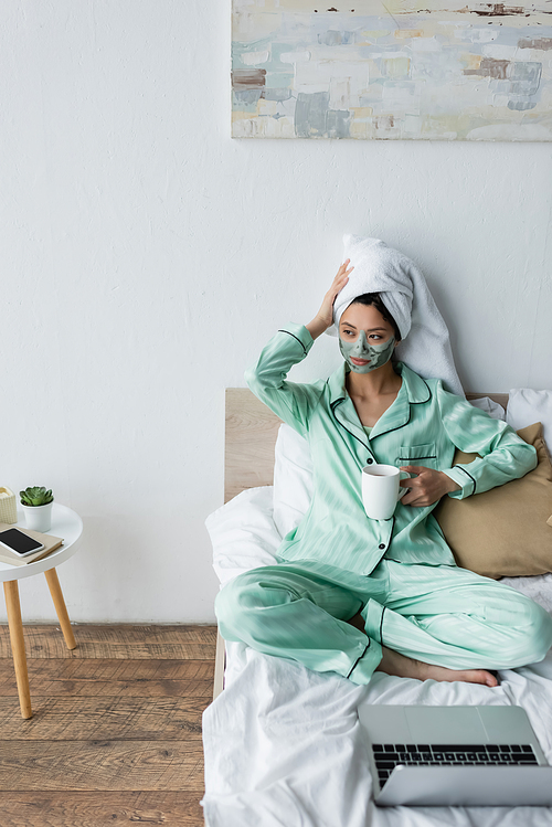 asian woman in clay mask and pajamas touching towel on head while sitting near laptop on bed