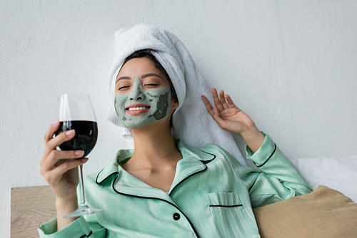 happy asian woman in pajamas, towel on head and clay mask holding red wine in bed