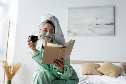 smiling asian woman in pajamas and clay mask holding red wine while reading book in bedroom