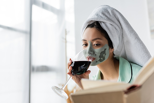 asian woman with clay mask and towel on head drinking red wine while reading book at home