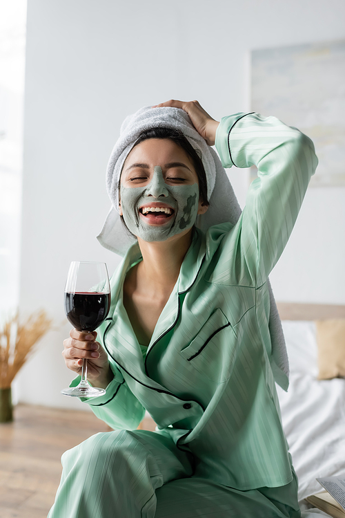 excited asian woman in clay mask and towel on head holding glass of red wine and laughing with closed eyes
