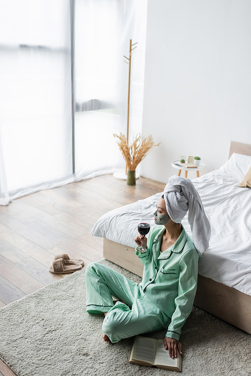 high angle view of asian woman in pajamas and clay mask sitting on floor in bedroom with glass of red wine