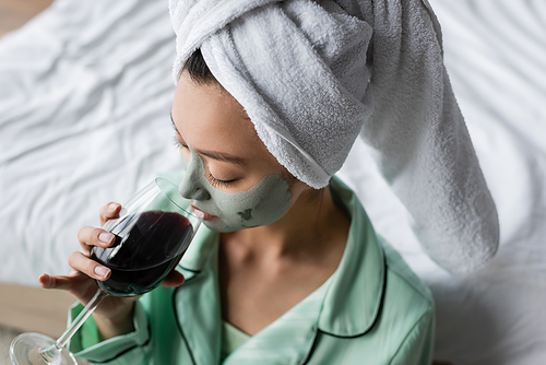 high angle view of asian woman in clay mask and towel on head drinking red wine in bedroom