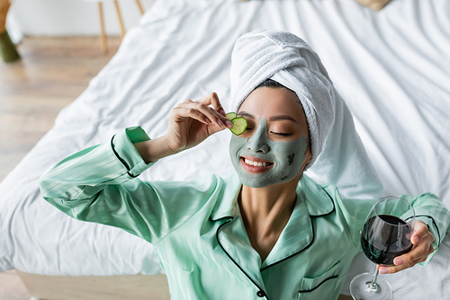 joyful asian woman in clay mask applying cucumber slices on eye while holding glass of red wine