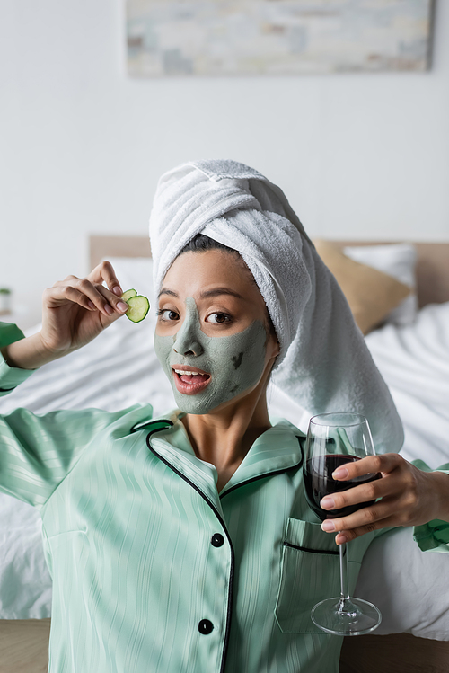 young asian woman in clay mask and pajamas holding cucumber slices and glass of red wine while 
