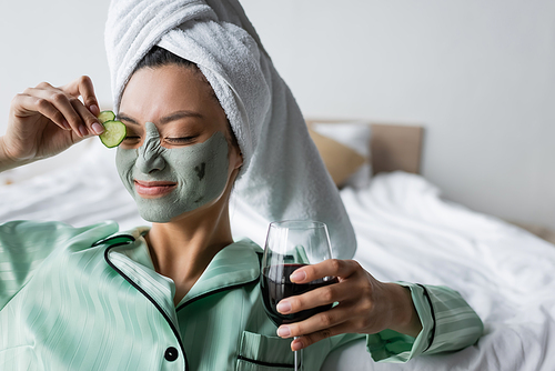 cheerful sian woman in clay mask applying cucumber slices on eye while holding glass of red wine