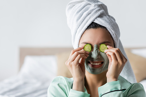 cheerful asian woman in clay mask frowning while applying cucumber slices on eyes