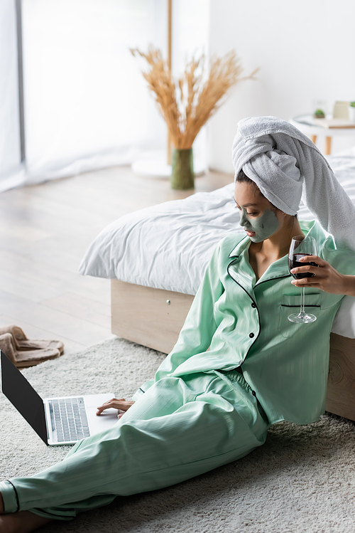 asian woman in clay mask and pajamas sitting on floor with glass of red wine and using laptop