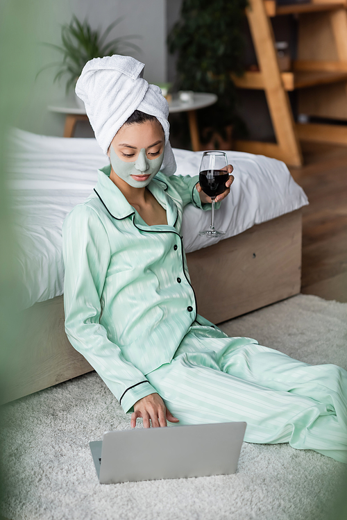 asian woman in pajamas and clay mask holding red wine and using laptop on floor in bedroom