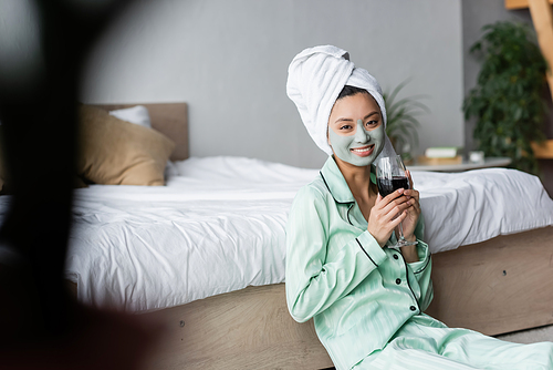 young asian woman in clay mask, pajamas and towel on head holding red wine in bedroom on blurred foreground