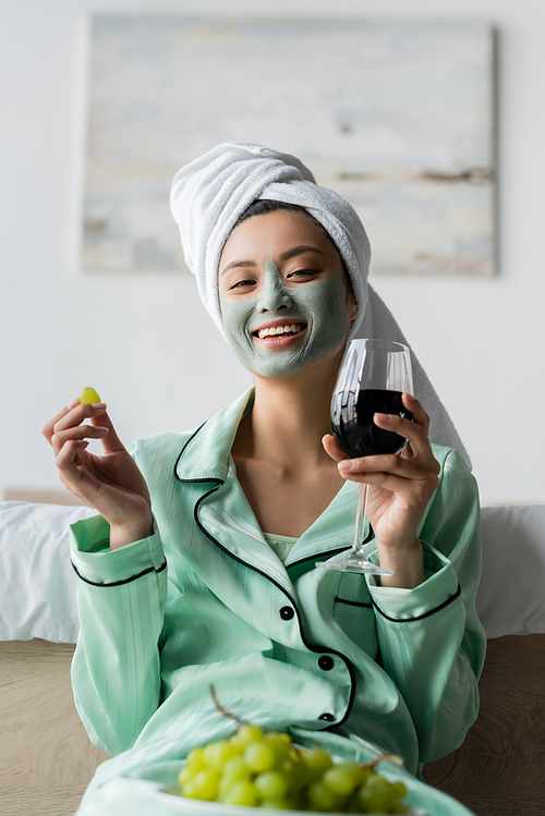 cheerful asian woman in clay mask and towel on head holding grape and red wine in bedroom