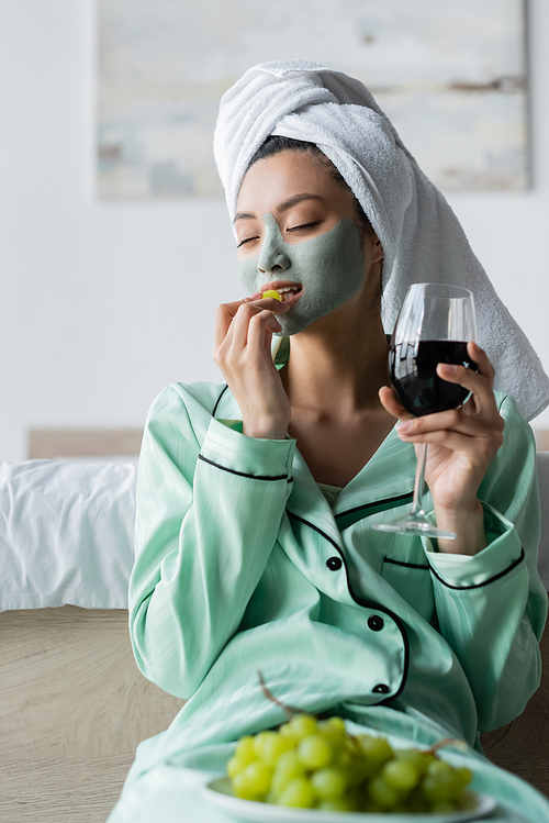 asian woman in towel on head and clay mask eating grape while holding glass of red wine