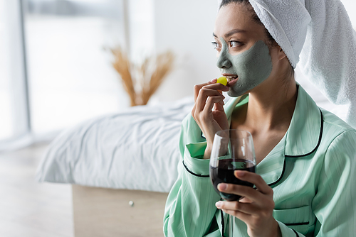 asian woman in clay mask eating grape while holding glass of red wine in bedroom