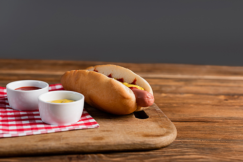 chopping board with hot dog, ketchup and mustard in bowls on wooden table isolated on grey