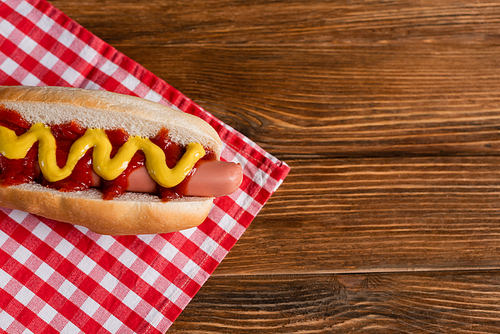 top view of tasty hot dog with sauces, and checkered napkin on wooden table