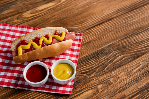 bowls with ketchup and mustard near tasty hot dog on plaid table napkin and wooden surface