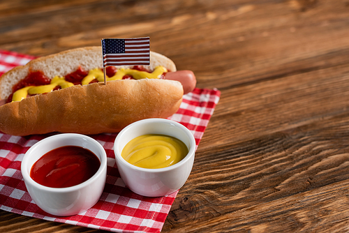 bowls with sauces near tasty hot dog with small american flag and plaid napkin on wooden table