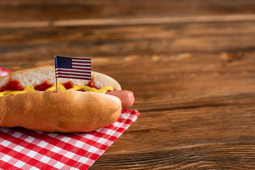 tasty hot dog with small american flag on plaid napkin and wooden table