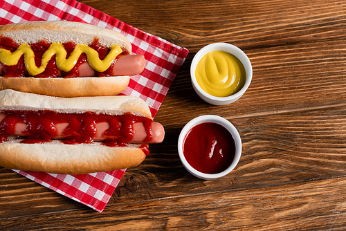 top view of hot dogs near sauce bowls and checkered napkin on wooden table