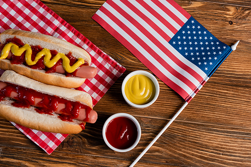 top view of small american flag near hot dogs, sauces and checkered table napkin on wooden surface