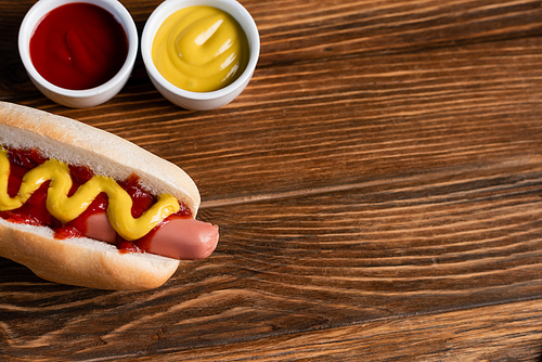 top view of mustard and ketchup in bowls near tasty hot dog on wooden surface