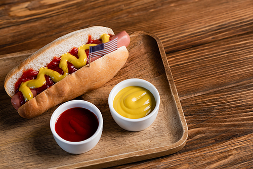 hot dog with small usa flag near bowls with sauces on wooden table