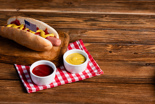 hot dog with small american flag on chopping board and sauces on plaid napkin and wooden table