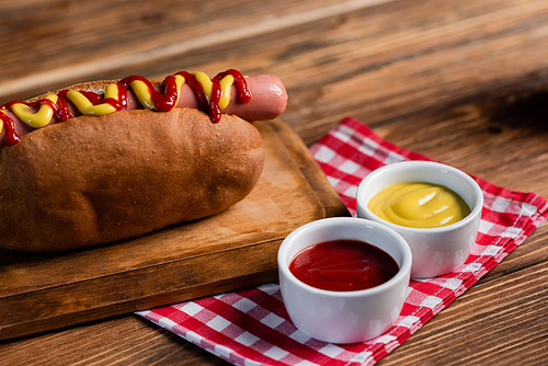 tasty hot dog on chopping board near bowls with mustard and ketchup and plaid napkin on wooden table