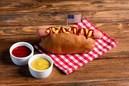 delicious hot dog with small usa flag near bowls with ketchup and mustard on wooden table