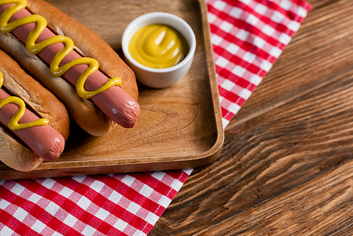 wooden tray with tasty hot dogs and bowl with mustard on checkered napkin and wooden table