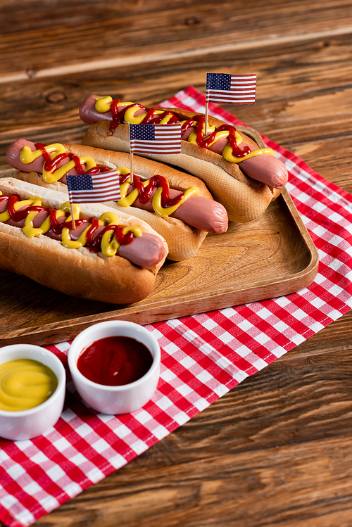 hot dogs with small usa flags on tray and plaid napkin near ketchup and mustard on wooden table